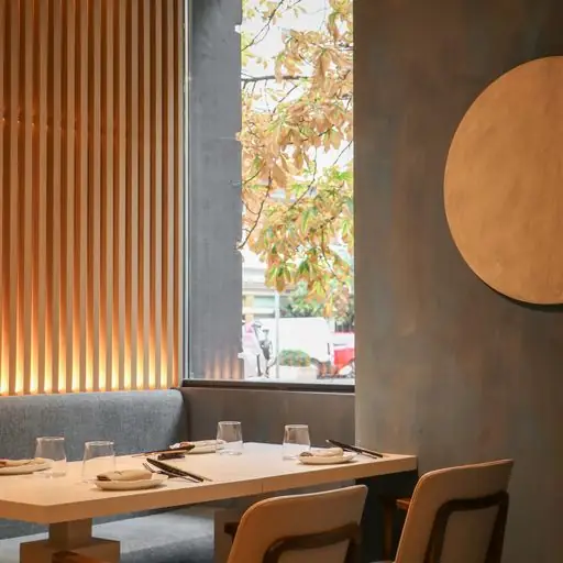 Restaurant interior with a grey banquette, pale wood table set with glasses and chopsticks, wooden slat wall and a large gold disc, a tree visible through the window