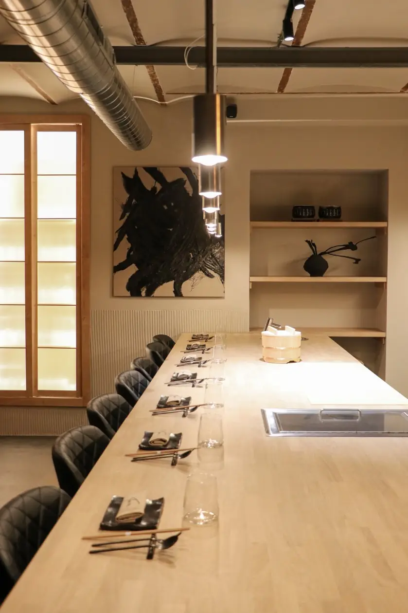 Long blond-wood sushi counter lined with black leather chairs and place settings of chopsticks and glasses, a black abstract painting and open wood shelving behind