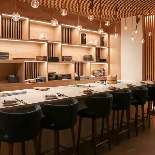 Blond-wood sushi bar counter with dark upholstered stools and wall shelving stacked with plates and ceramics under warm pendant lighting