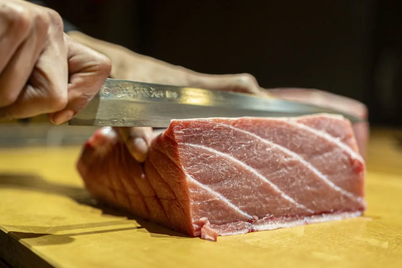 Hands slicing a fatty block of otoro tuna with a long Japanese knife on a yellow cutting board