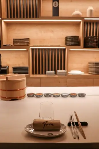 Sushi counter place setting with a glass, chopsticks and a small dish on a white plate, a row of condiment bowls and stacked plates on shelving behind