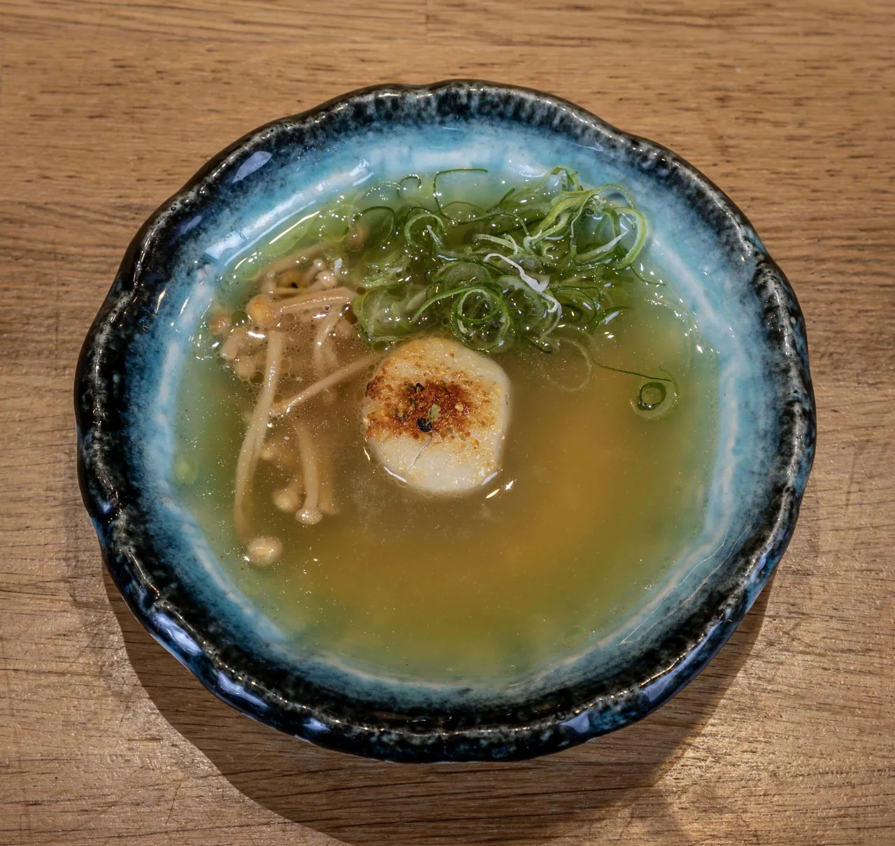 A blue-rimmed bowl of clear broth with mushrooms, a round pale piece and chopped green onion