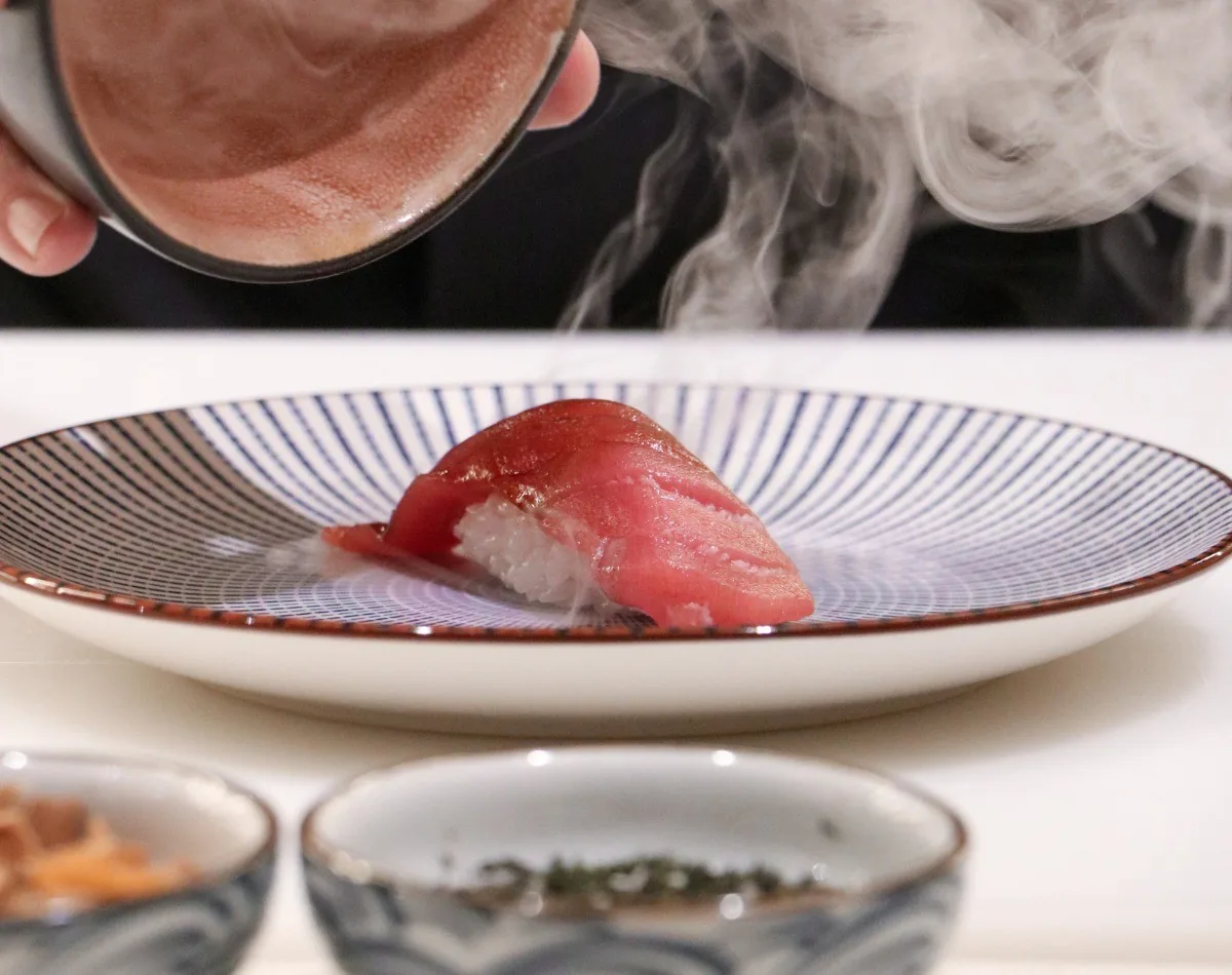 A single nigiri of red fish on a striped white plate with rising steam