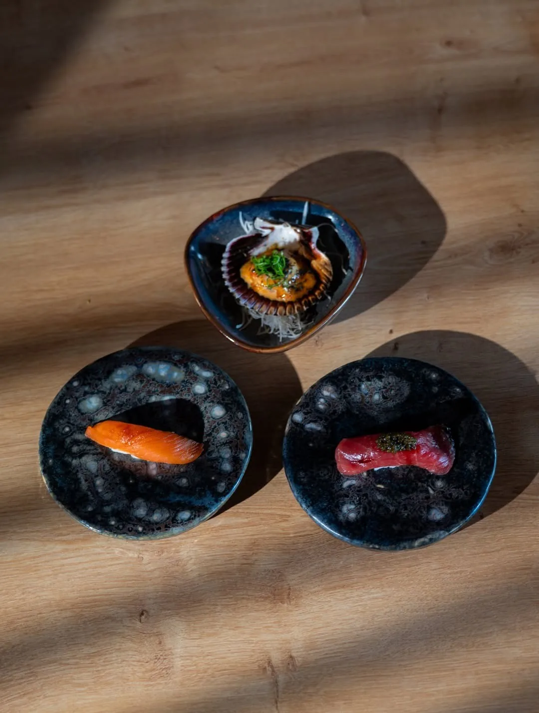 Three nigiri on small dark round dishes on a wooden table