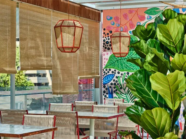 Sunlit terrace dining area with bamboo roll-up blinds, red-framed rattan lantern pendants, a colourful botanical mural and rope-woven chairs at tile-topped tables