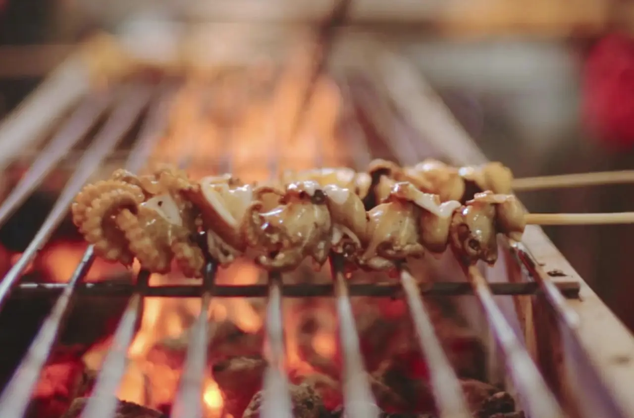A skewer of glazed octopus and squid pieces cooking over open flames and glowing charcoal on a steel grill bar
