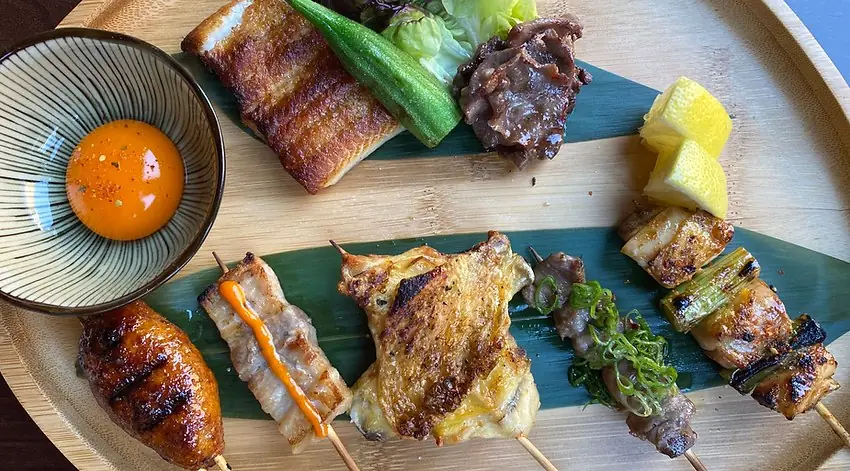 A round wooden tray lined with green leaves holding grilled chicken and pork skewers, a glazed chicken wing, a piece of eel, okra and a bowl of raw egg yolk with lemon wedges