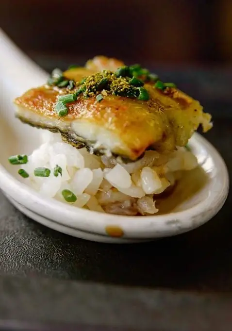 A white ceramic spoon holding a piece of glazed grilled eel over rice, scattered with chopped chives and green powder