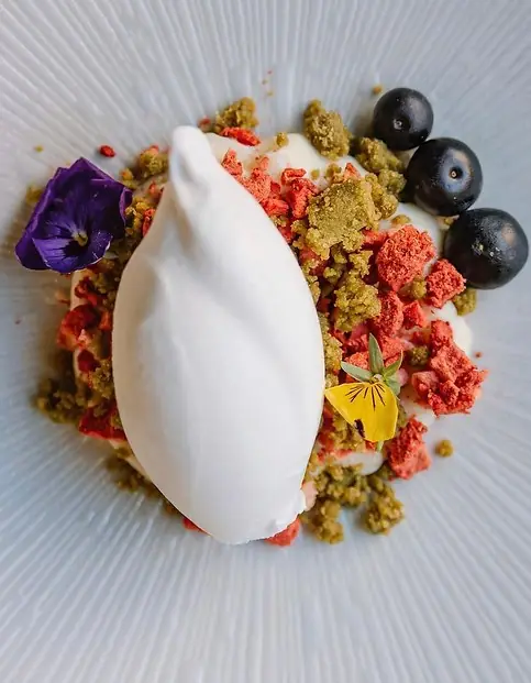 A white quenelle of cream on a ridged plate with strawberry crumble, green crumb, dark grapes and purple and yellow pansies