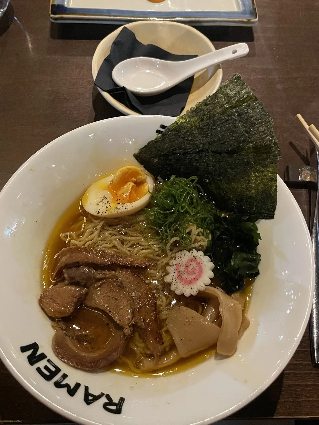 A white bowl marked RAMEN with noodles in broth, sliced pork, half a soft-boiled egg, nori, seaweed and a fish cake