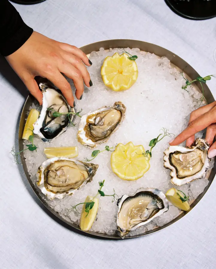 Six freshly shucked oysters on ice with lemon wedges and pea shoots on a round metal platter, being served at Altamar