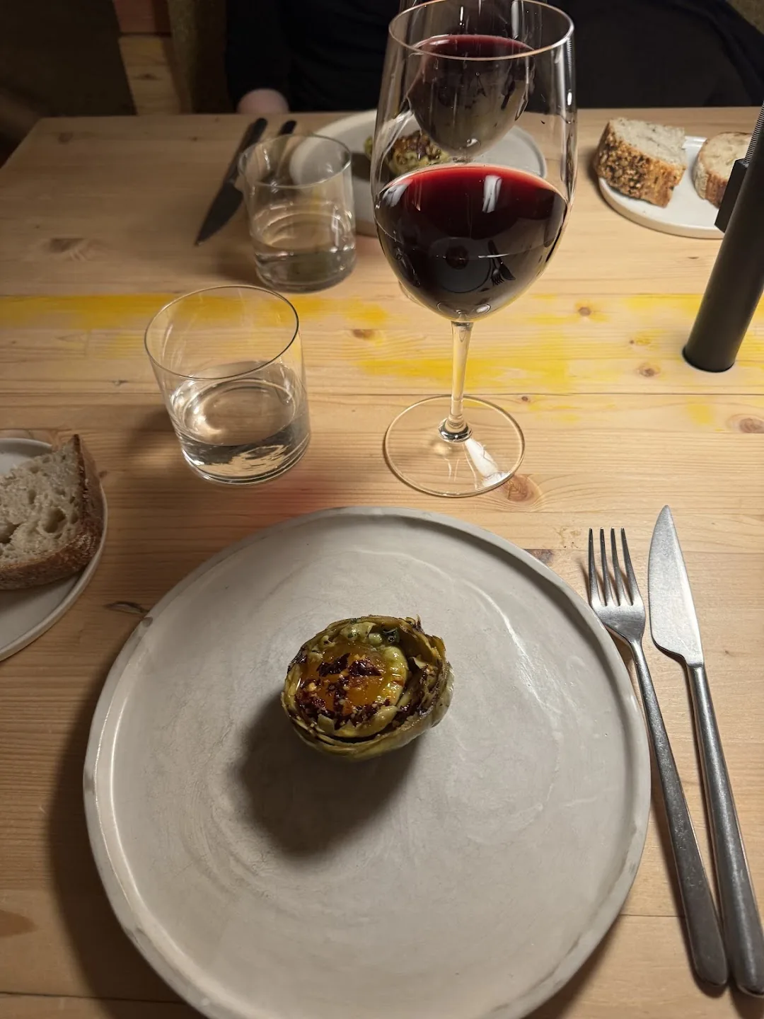 A halved grilled artichoke topped with egg yolk and chili flakes, served with a glass of red wine and bread