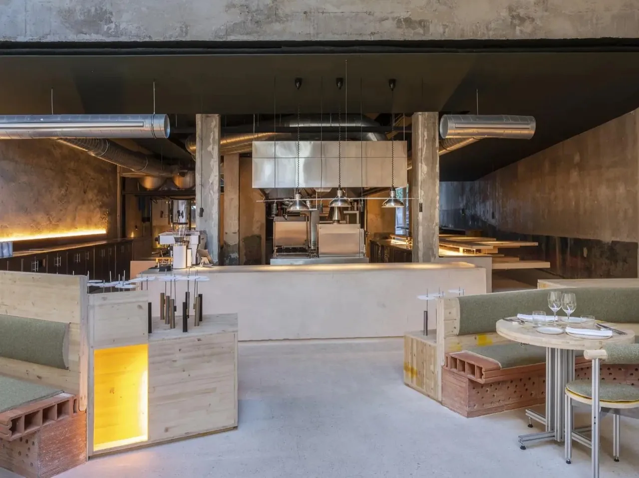 A dining hall with an open kitchen in the middle, green banquettes and round tables