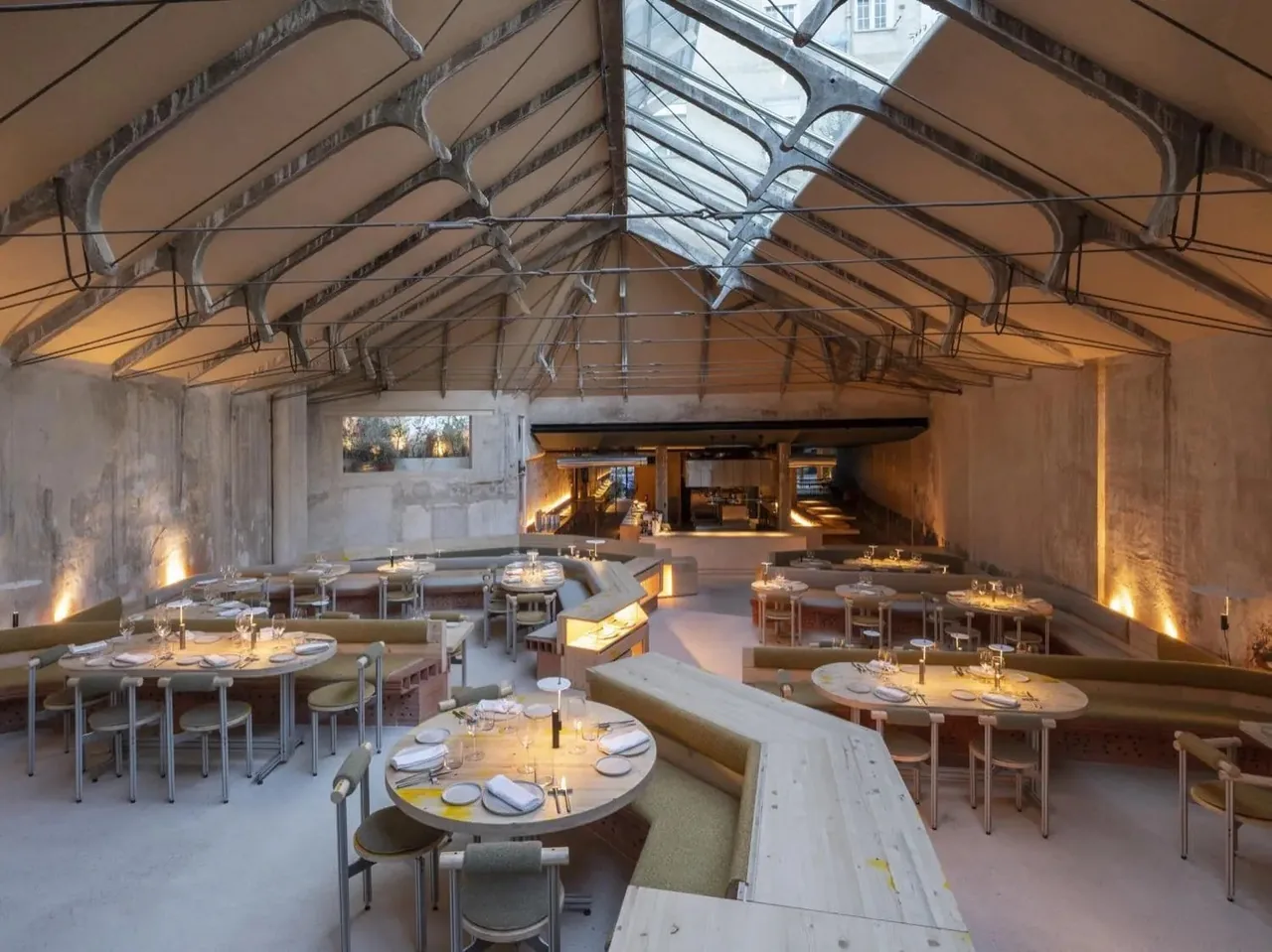 A large industrial dining hall with a glass roof, round tables and warm lit counters