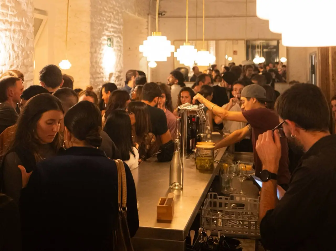 Crowded whitewashed-brick bar interior with pendant lights, patrons packed along a steel bar counter and a bartender pouring behind it