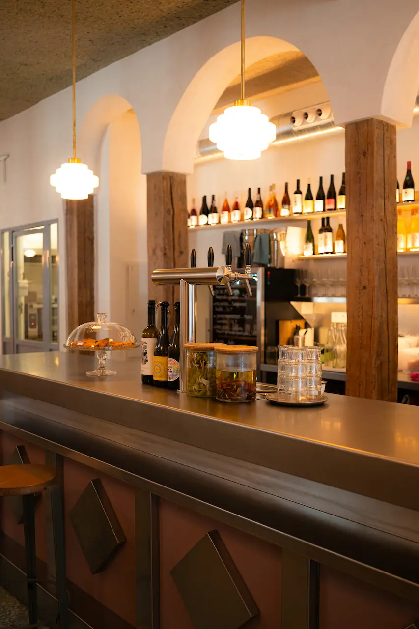 Steel bar counter with beer taps, a cake dome and infused jars, under whitewashed arches and glass pendant lamps with a lit bottle shelf behind