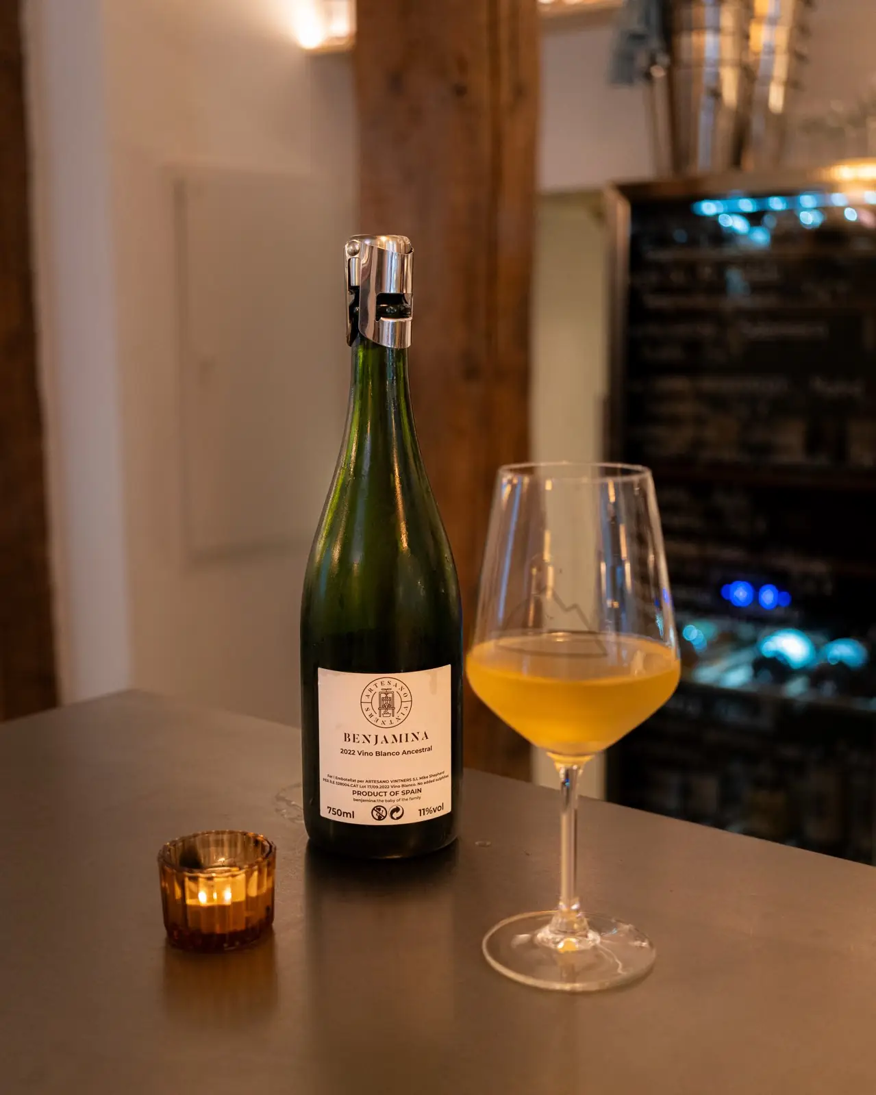 Glass of cloudy orange natural wine beside an opened green bottle labeled Benjamina and a lit candle on a steel bar top