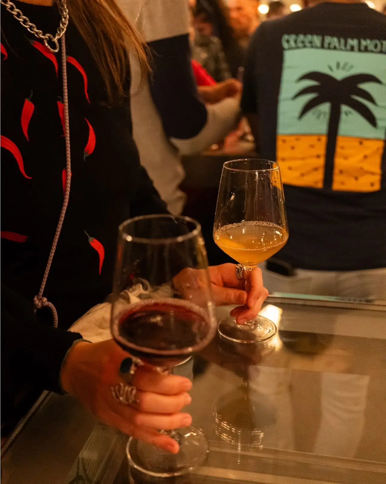 Two hands holding stemmed glasses of red and orange wine over a glass-topped table in a busy bar