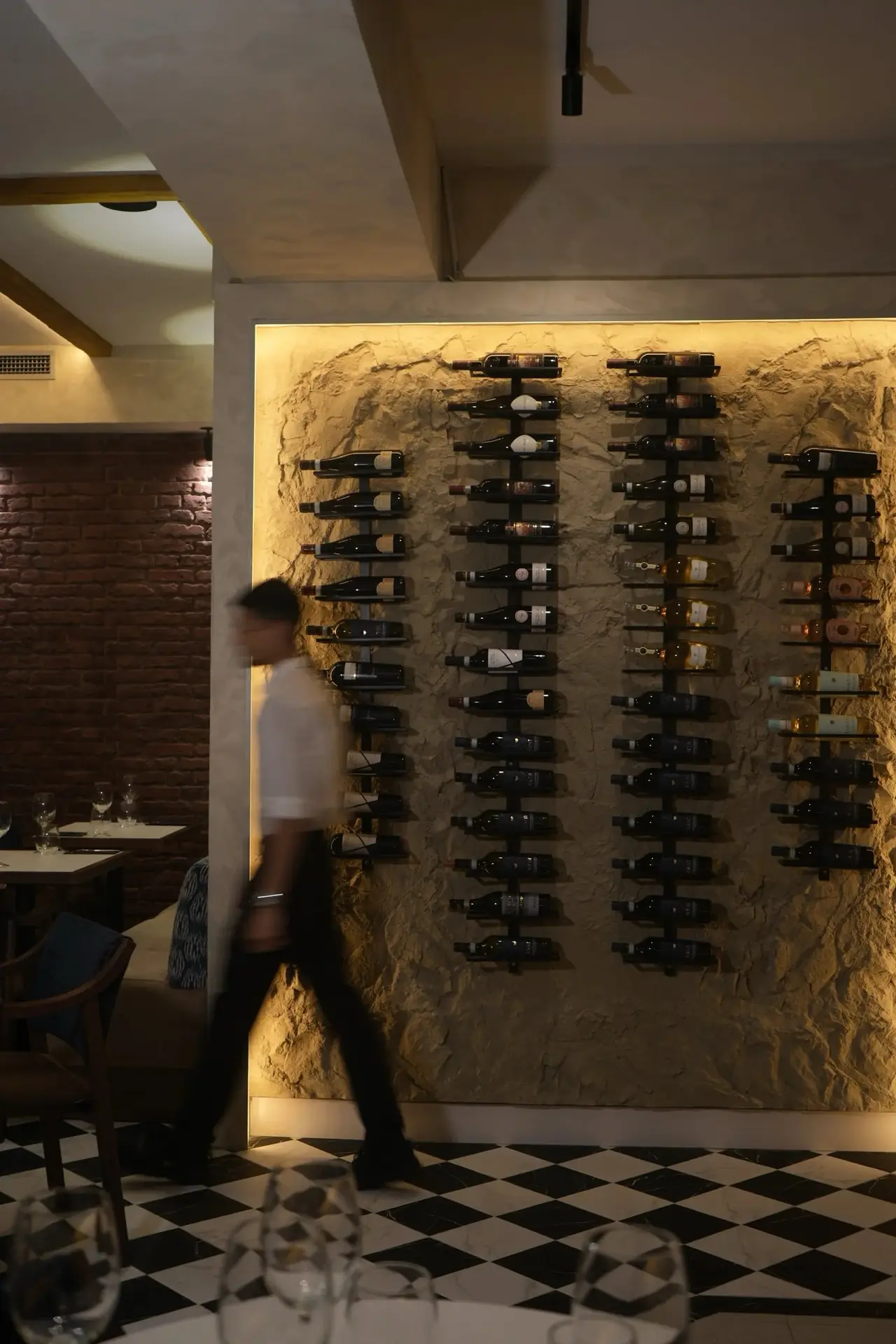 Dining room with a rock-textured feature wall of backlit wine bottles on black racks, exposed brick, black-and-white checkerboard tile floor, and a waiter walking past in motion blur