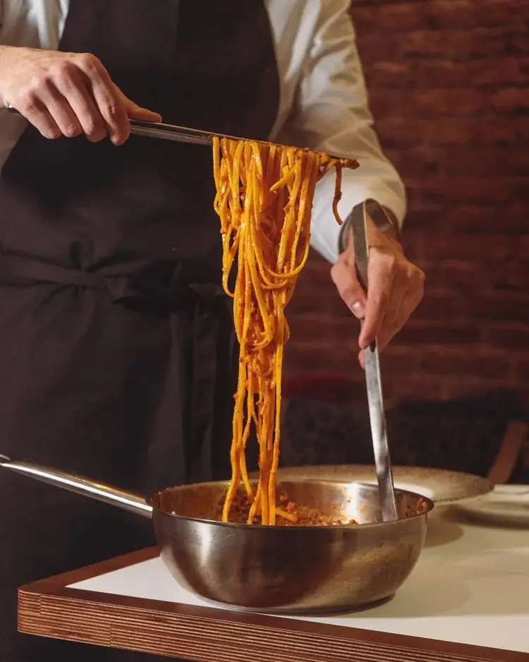 Hands in a black apron lifting a portion of tomato-sauced tonnarelli pasta with tongs from a copper pan set on a wooden table