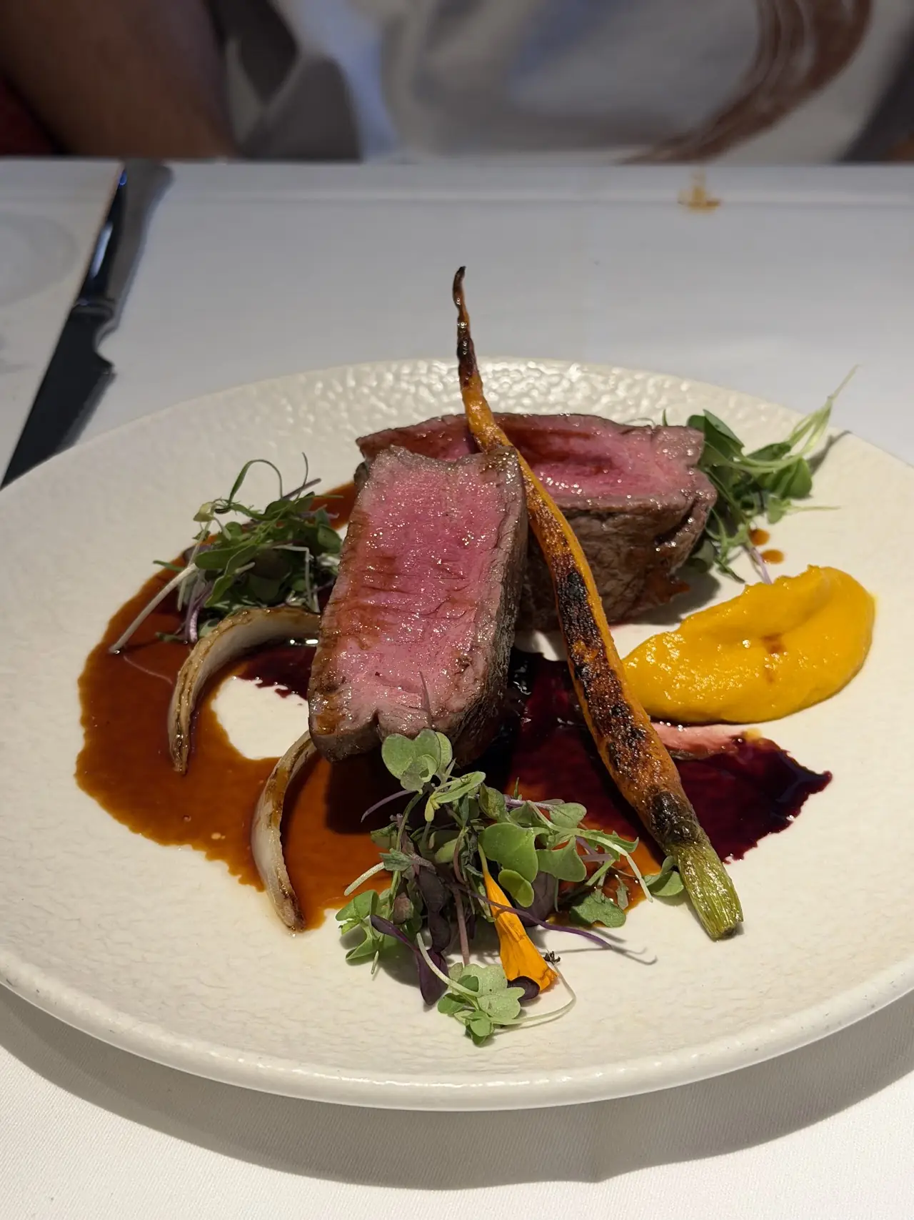 Sliced pink beef with grilled carrots, greens, and an orange puree on a white plate.