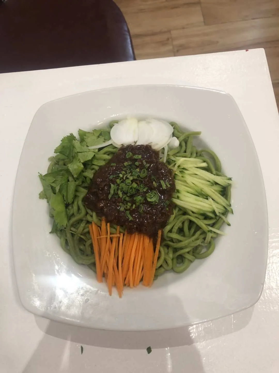 A white plate of noodles topped with shredded carrots, cucumber, and a scoop of dark sauce