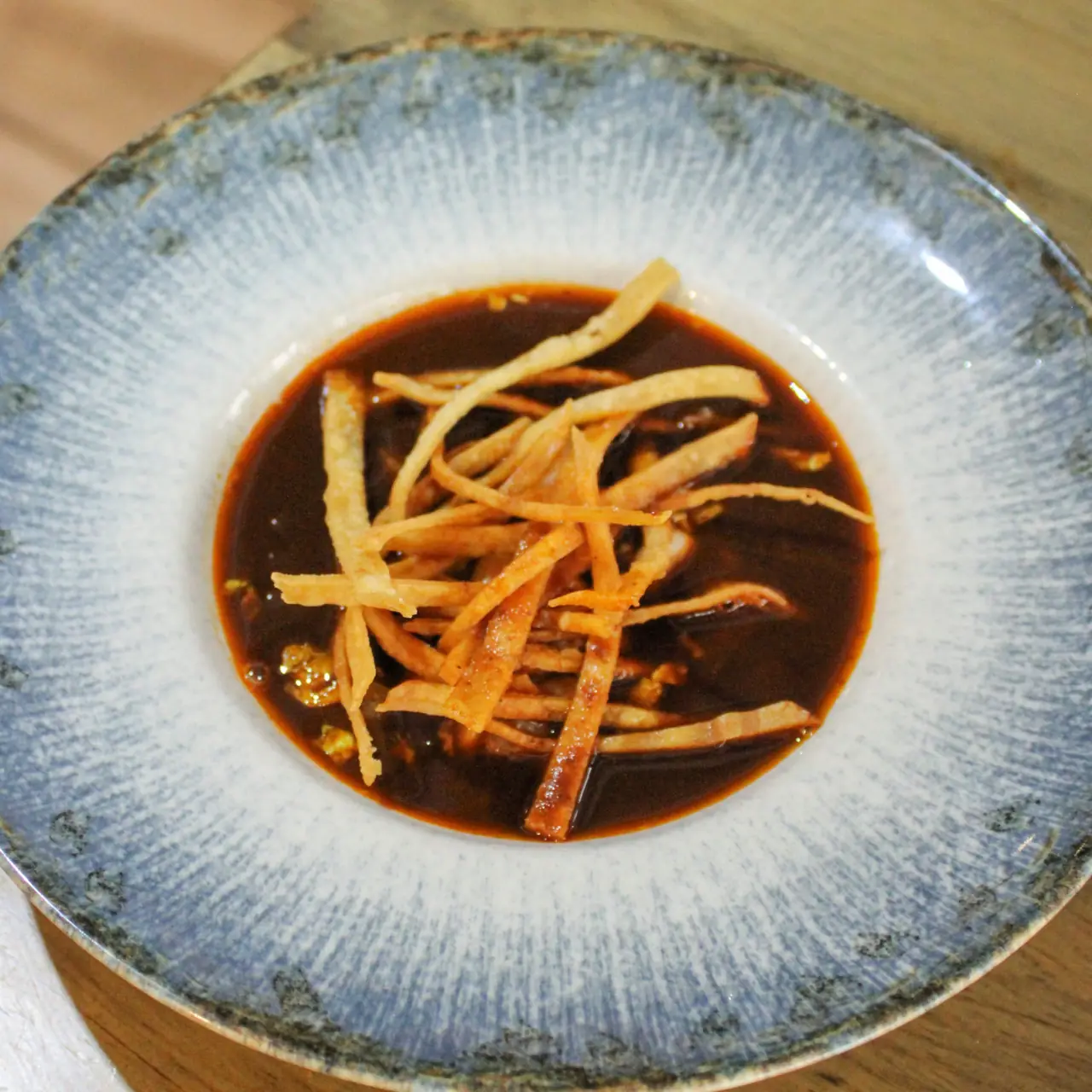 A shallow blue speckled ceramic bowl of dark reddish-brown broth topped with a tangle of crisp fried tortilla strips, on a pale wooden table
