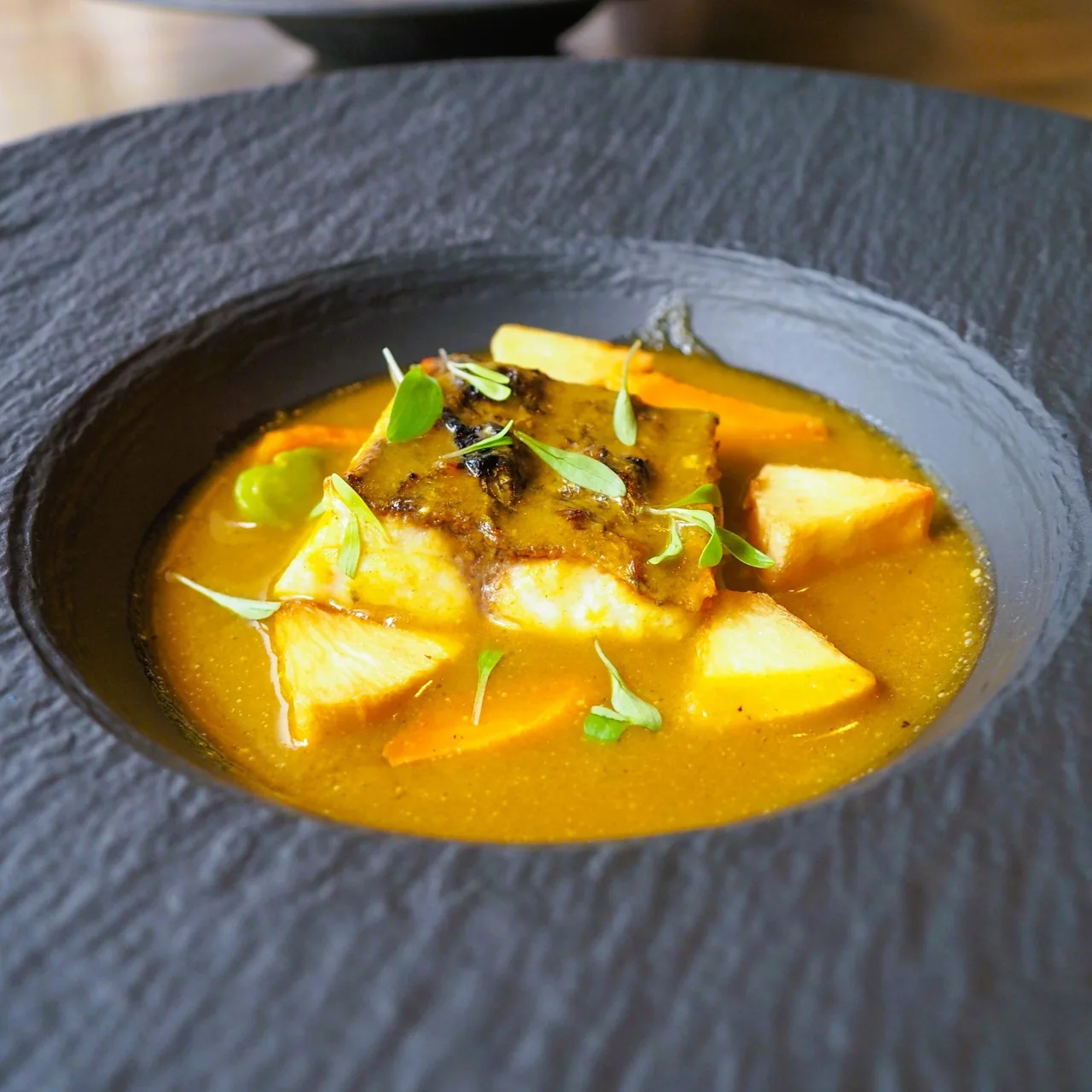 A dark bowl of yellow broth with pieces of potato and vegetables