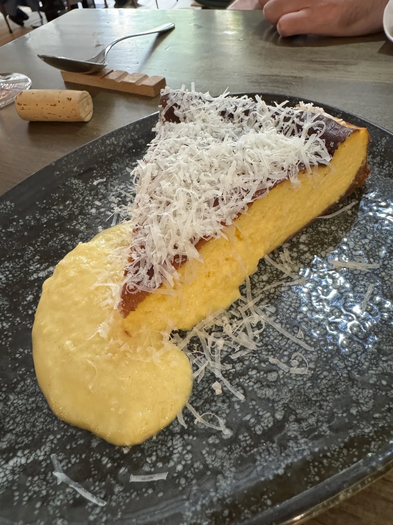 A slice of baked cheesecake topped with grated cheese on a dark speckled plate with a smear of yellow cream