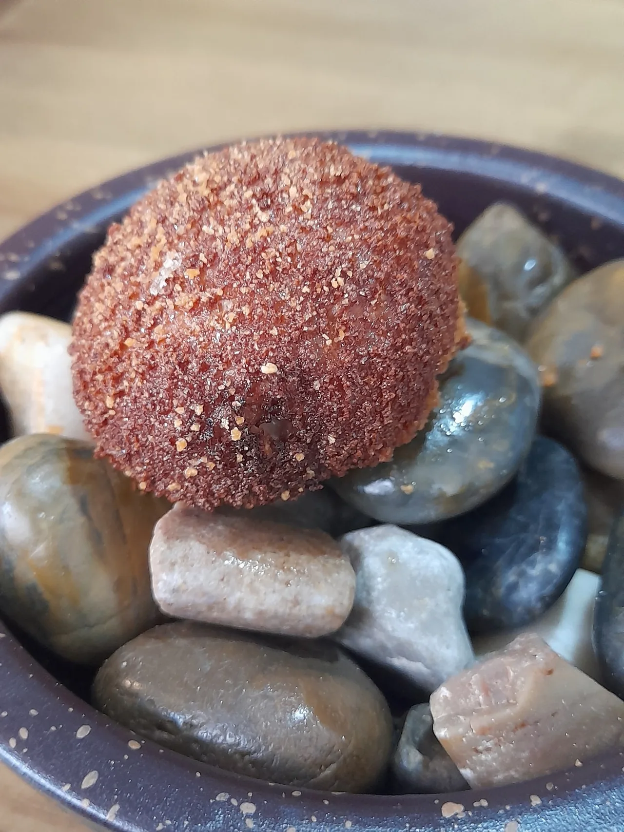 A single round breadcrumbed fried croqueta sitting on pebbles in a dark bowl