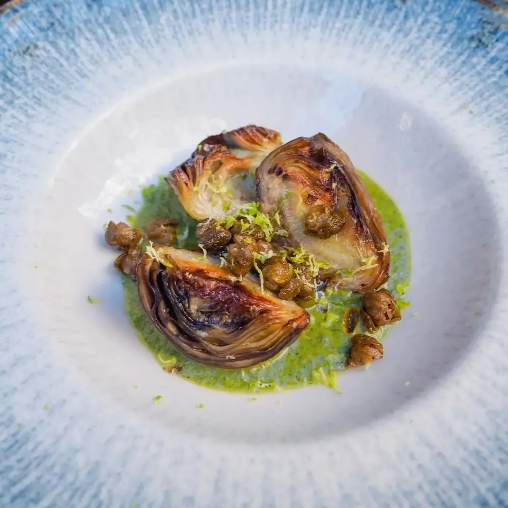 Charred artichoke halves with crispy capers on a green herb sauce, white bowl