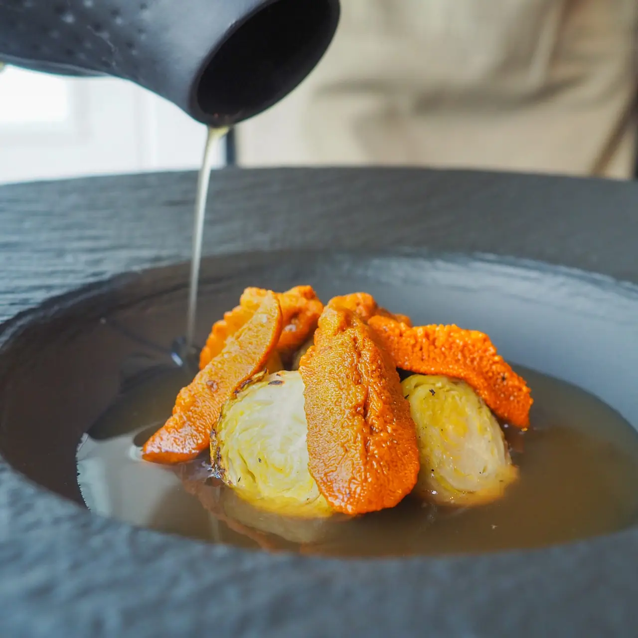 Broth poured over orange sea-urchin-like strips and charred baby cabbages on a dark plate