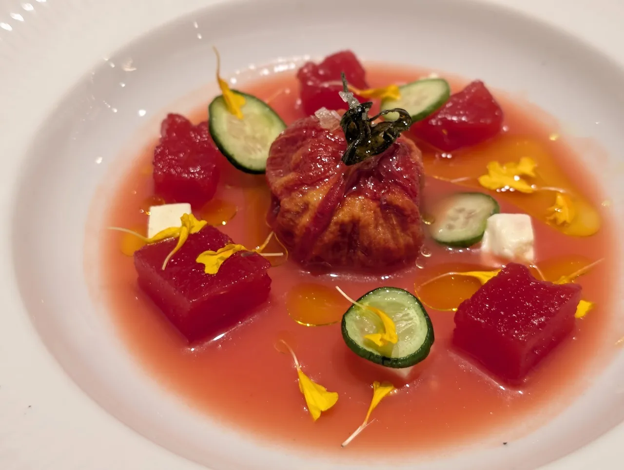 A plate of diced red tomato and a stuffed parcel in a pink broth with cucumber and yellow petals