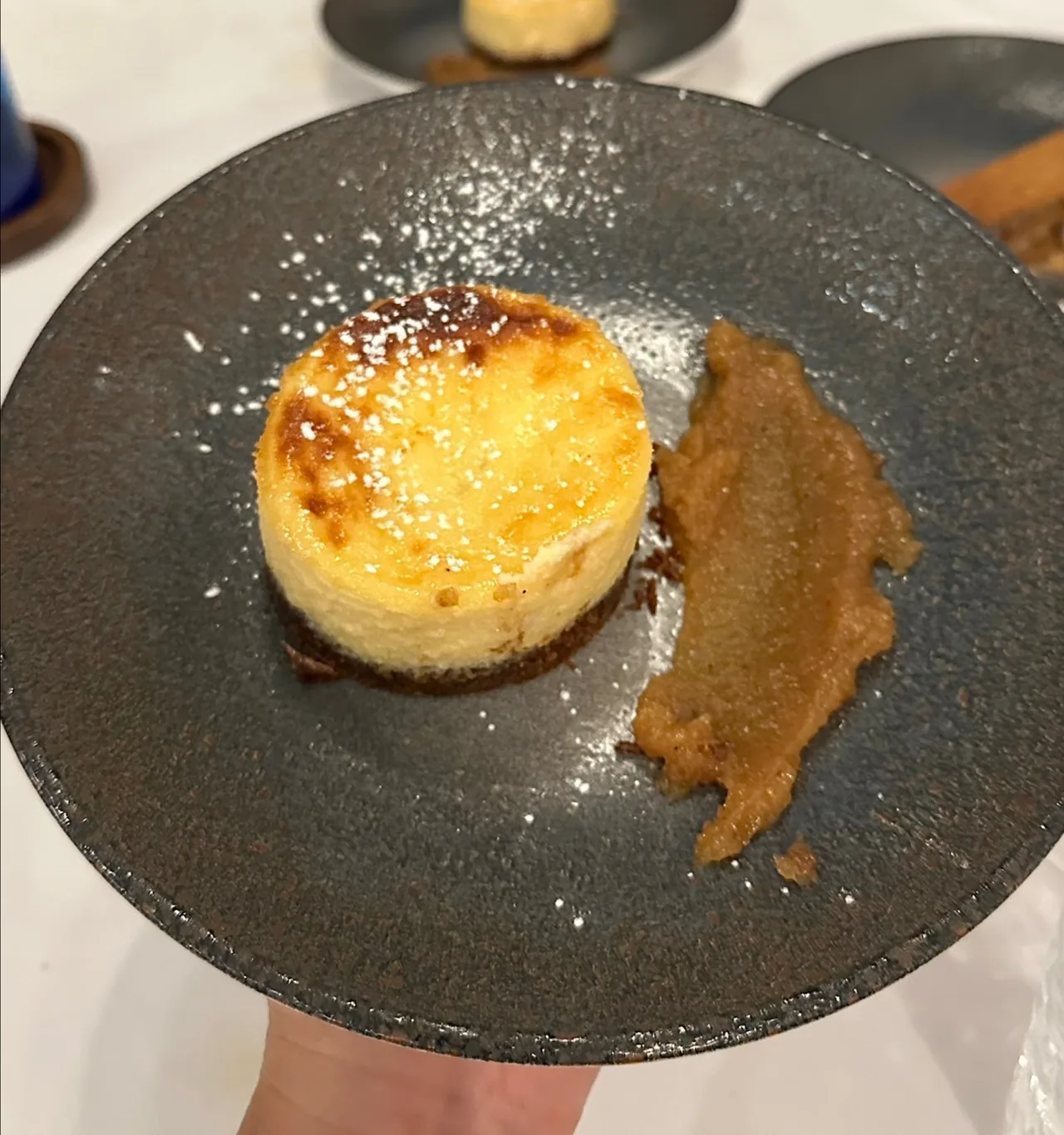 A round cheesecake with a browned top dusted with icing sugar on a dark plate beside a smear of brown puree