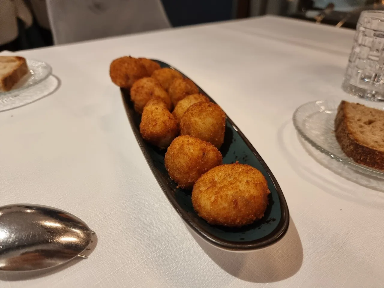 A dark oval plate of round golden croquetas on a white tablecloth