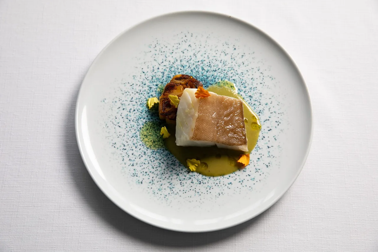 A piece of white fish with skin on a white plate speckled with blue dots and green sauce