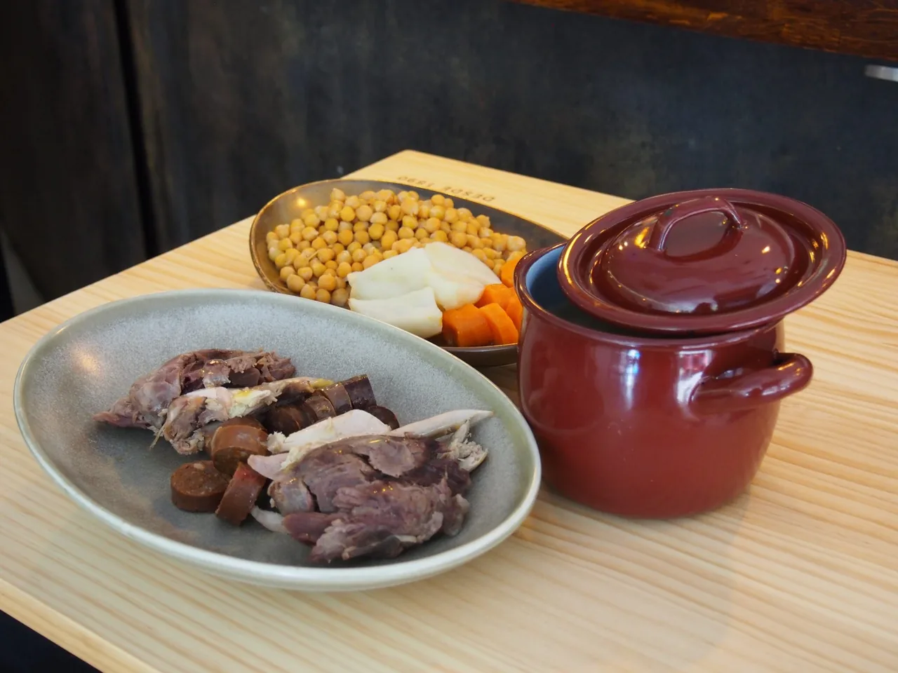 A grey plate of sliced meat next to a red lidded pot and a bowl of chickpeas on a wooden table