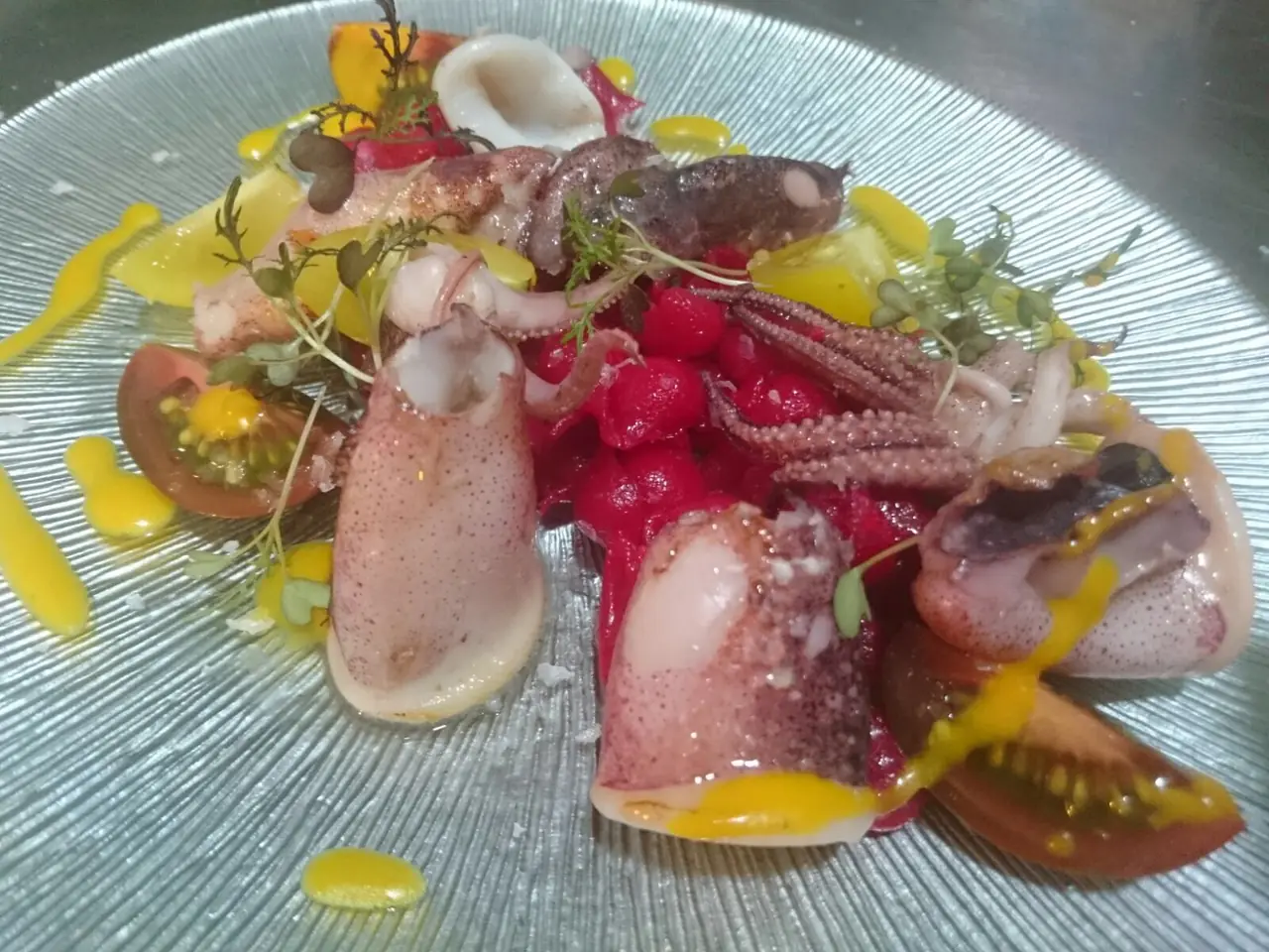 Plate of grilled baby squid with tomato wedges, red berry garnish and herbs, yellow sauce drizzle