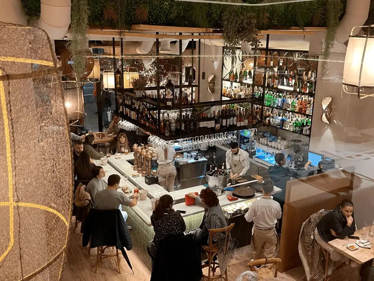 Overhead view of a busy central marble bar with a bartender, guests on stools, hanging plants and tall shelves stacked with wine and spirit bottles
