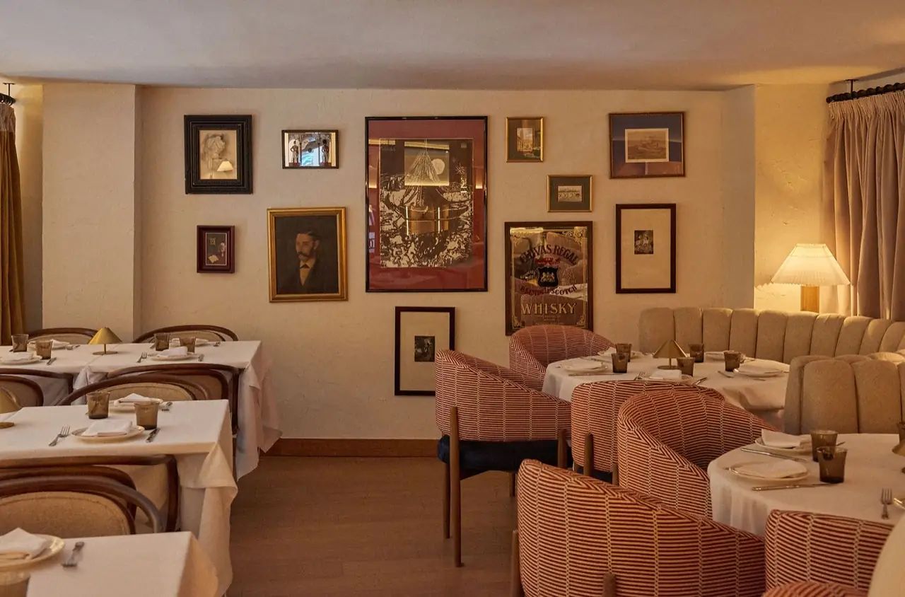 Warm-toned dining room with white-clothed tables, striped and cream upholstered chairs, and a wall hung with framed vintage art and a Chivas Regal whisky mirror