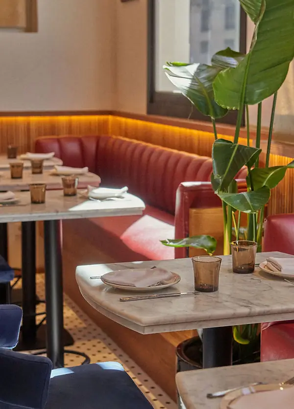 Marble-topped tables set with plates and amber glasses beside a red leather banquette with a large leafy plant