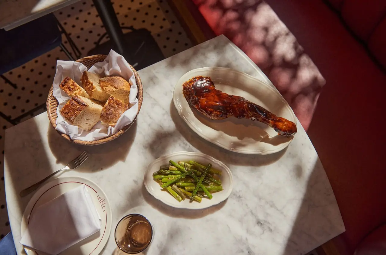 Glazed roast chicken leg on an oval plate with a side dish of grilled asparagus and a basket of bread on a marble table