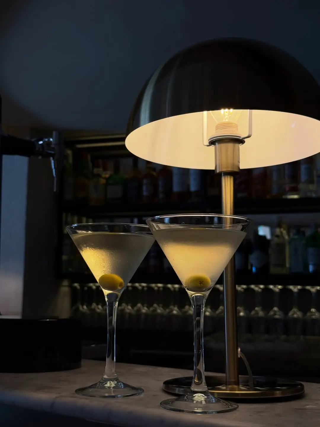 Two martini glasses with olive garnish on a bar counter beneath a warm brass lamp