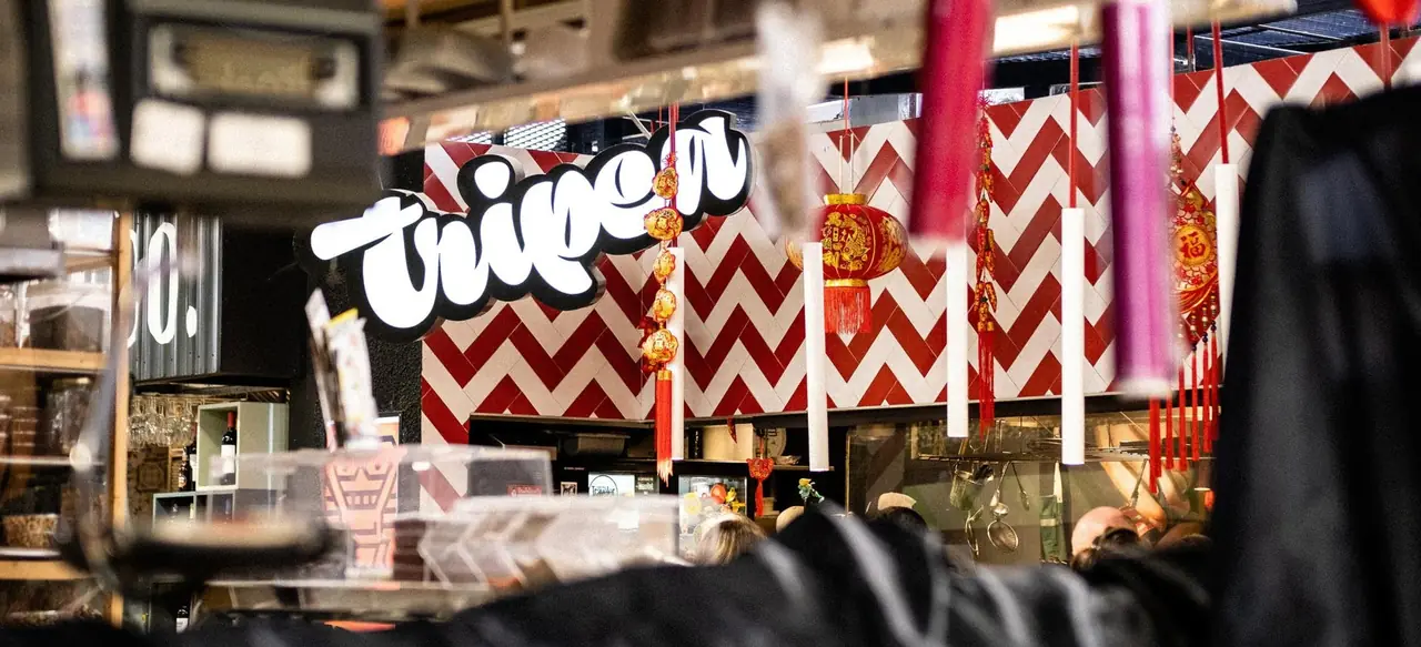 The Tripea stall inside a market hall, its illuminated white script sign above a red-and-white chevron tiled wall hung with red Chinese lanterns and tassels
