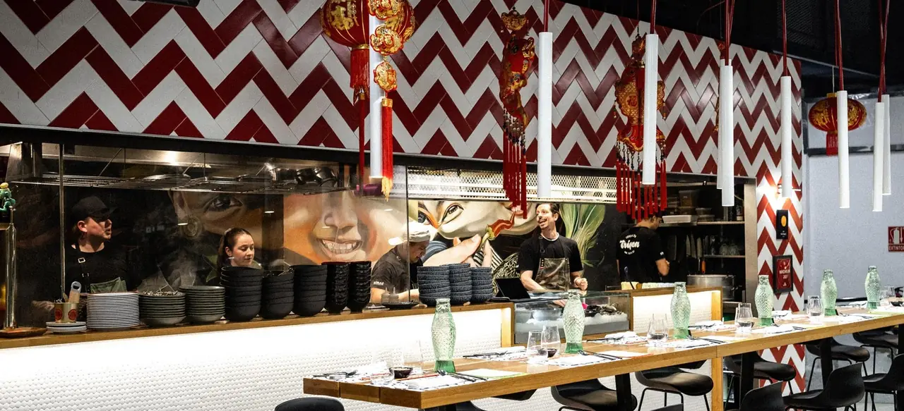 The open kitchen counter with cooks working behind glass, stacks of black bowls and white plates, a long wooden bar laid with place settings and green fish-shaped water bottles under red-and-white chevron tiles