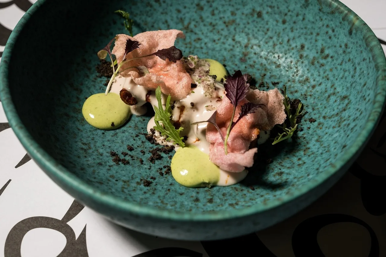 A green speckled bowl with slices of cured fish, a yellow sauce and small leaves