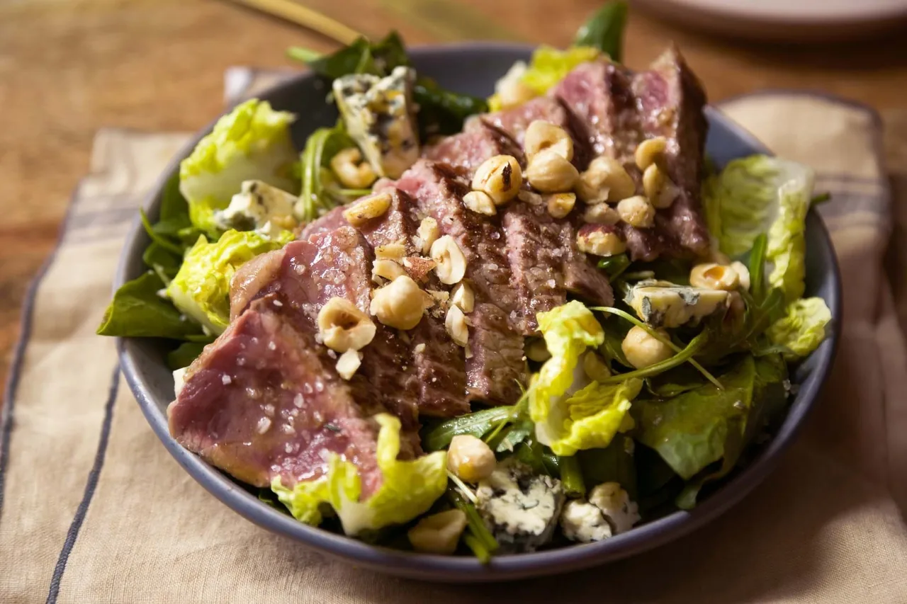 A dark bowl of salad with sliced steak and nuts