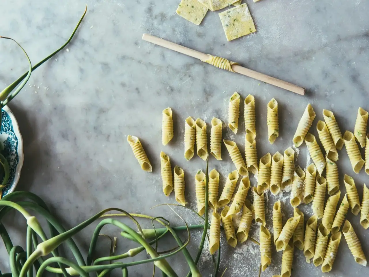 Fresh handmade garganelli pasta being rolled with a ridged board and dowel on a flour-dusted marble surface, with garlic scapes alongside