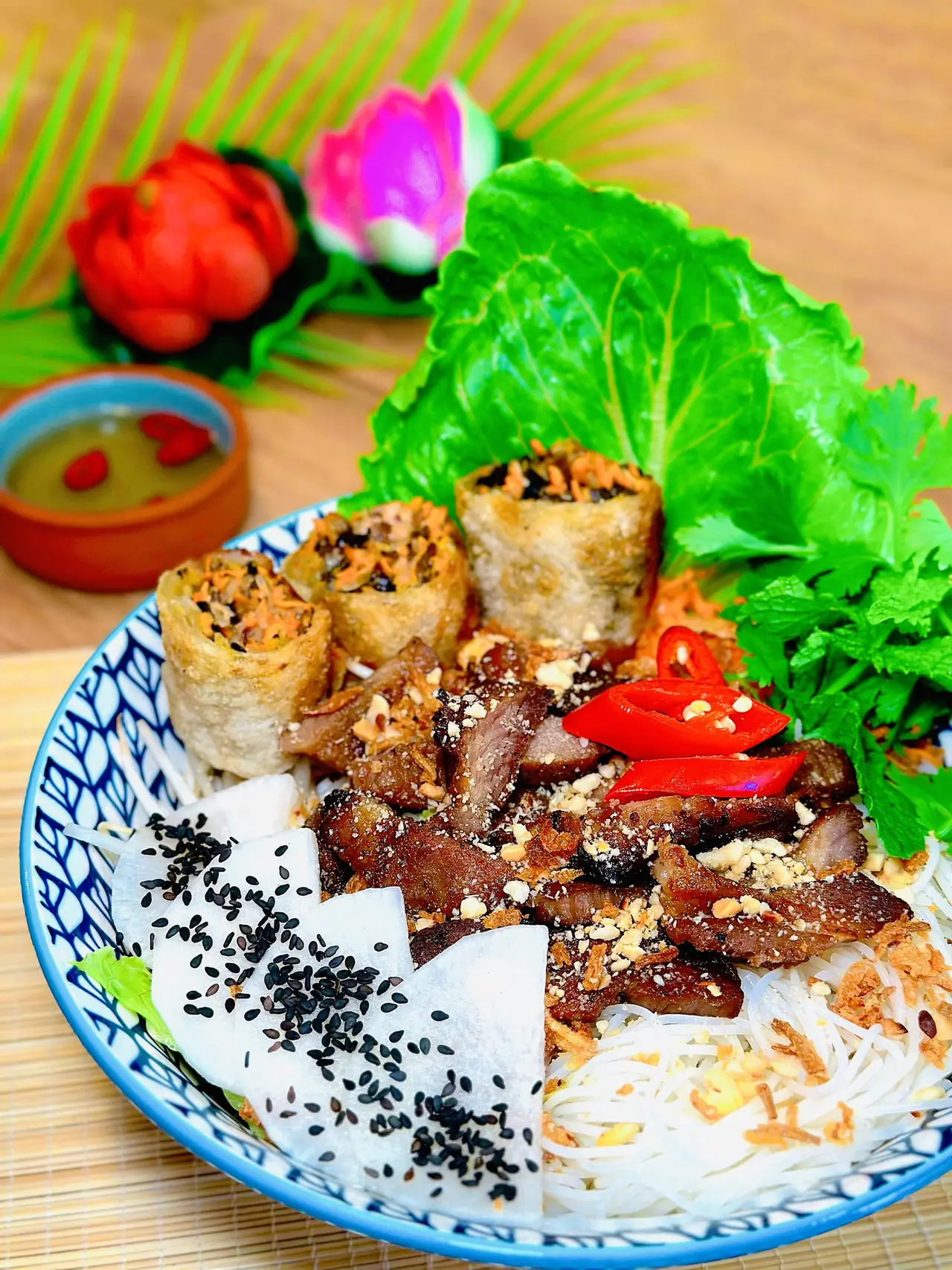 Bowl of rice vermicelli topped with sliced grilled pork, crispy fried rolls, black sesame, daikon, crushed peanuts, and fresh herbs with a dipping sauce, Tutu Vietnamese Cuisine