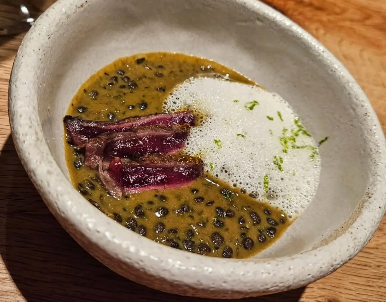 Seared rare meat slices over lentil sauce with citrus foam and zest in ceramic bowl